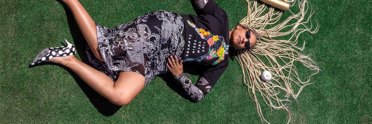a Black woman in chic clothes, polka dot heels, and long white dreds laying on a baseball field