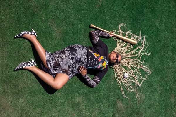 a Black woman in chic clothes, polka dot heels, and long white dreds laying on a baseball field