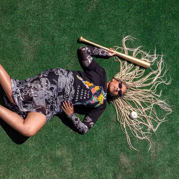a Black woman in chic clothes, polka dot heels, and long white dreds laying on a baseball field