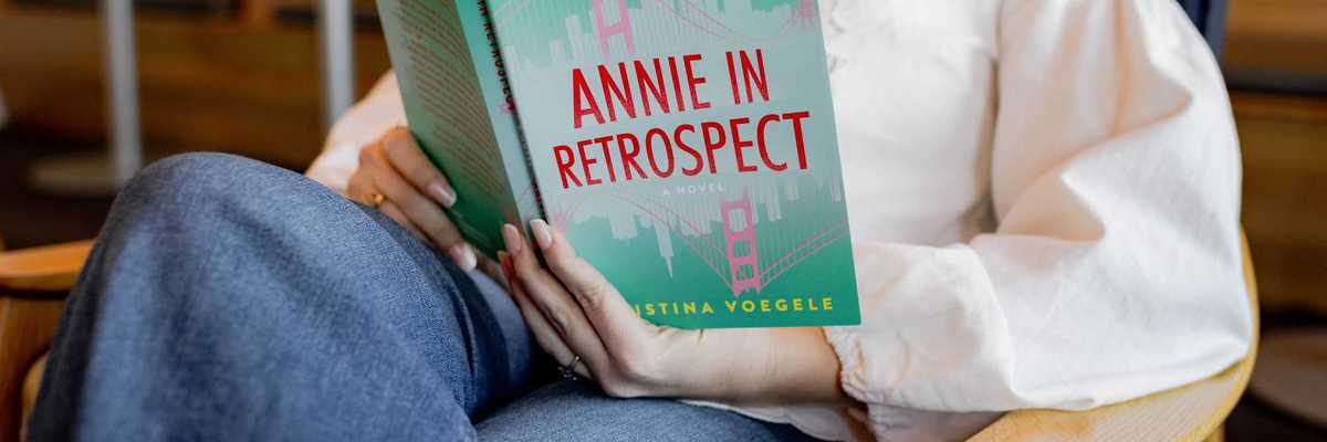 a blonde woman in a white shirt holding the book 'Annie In Retrospect'