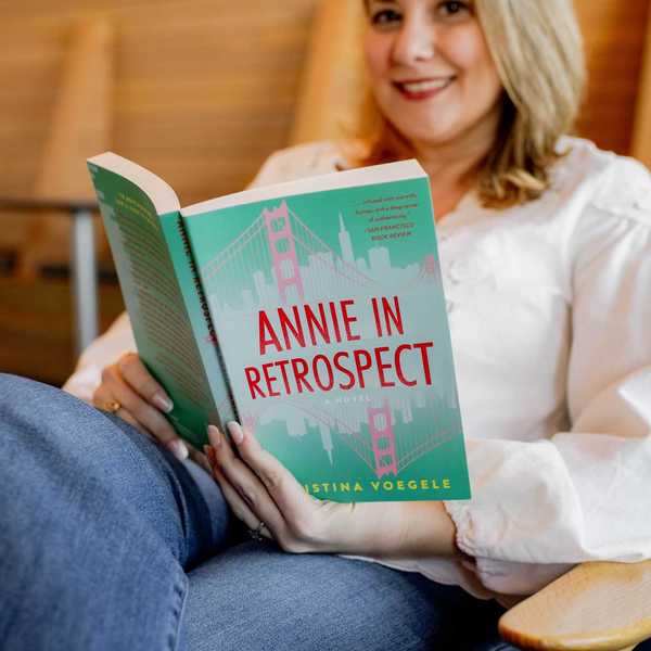 a blonde woman in a white shirt holding the book 'Annie In Retrospect'