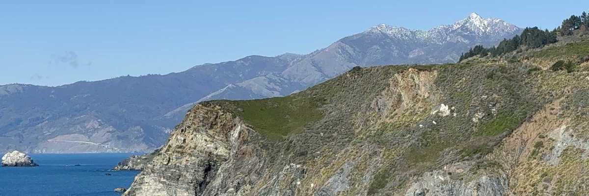 a cliff sticking out into the ocean with a mountain with snow in the background