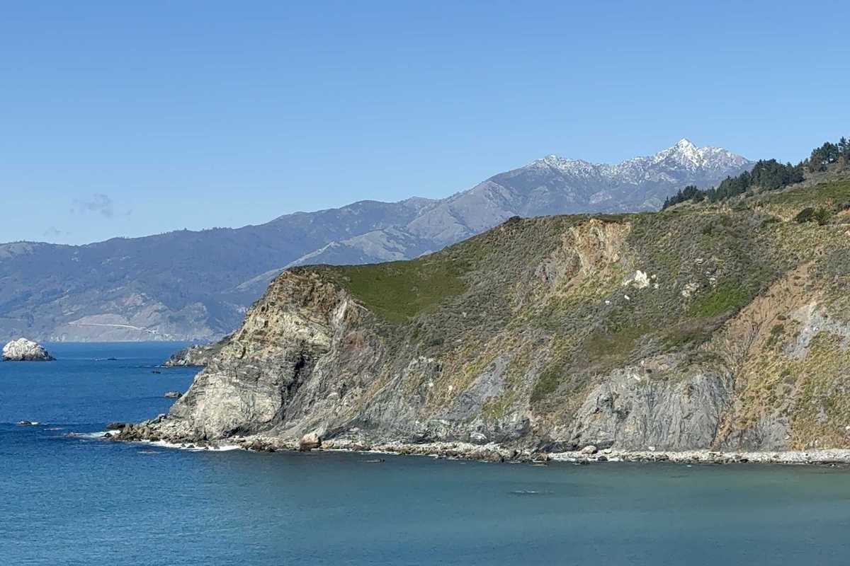 a cliff sticking out into the ocean with a mountain with snow in the background