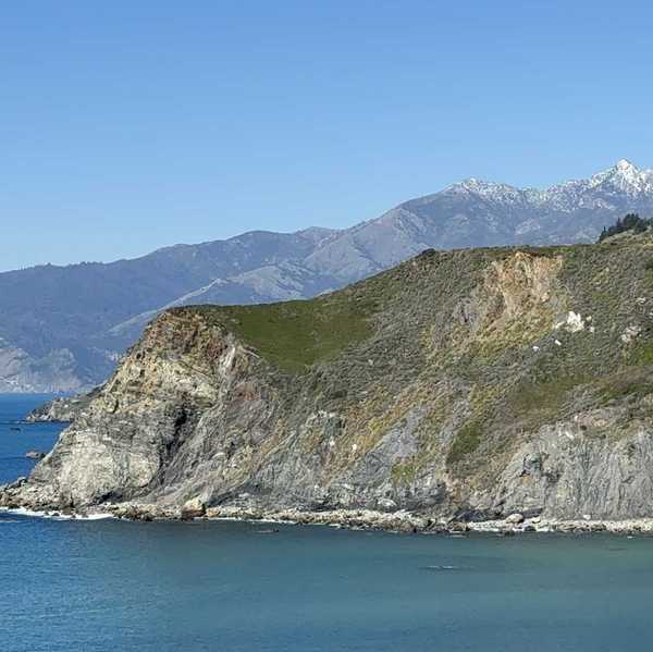 a cliff sticking out into the ocean with a mountain with snow in the background