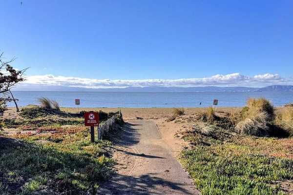 a dirt path leading to a sand beach with a blue body of water beyond