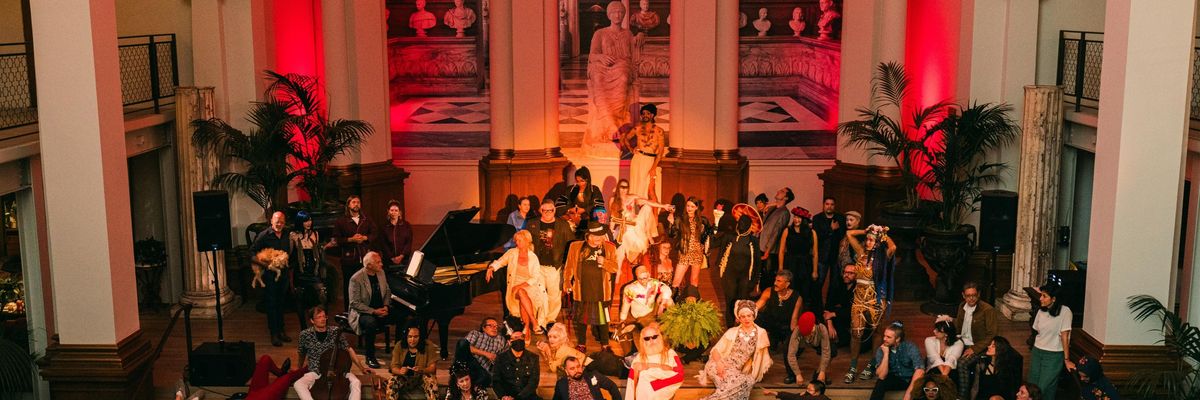 A diverse group of people in colorful attire gather in an ornate hall with red lighting.