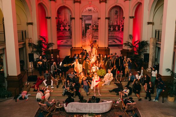 A diverse group of people in colorful attire gather in an ornate hall with red lighting.
