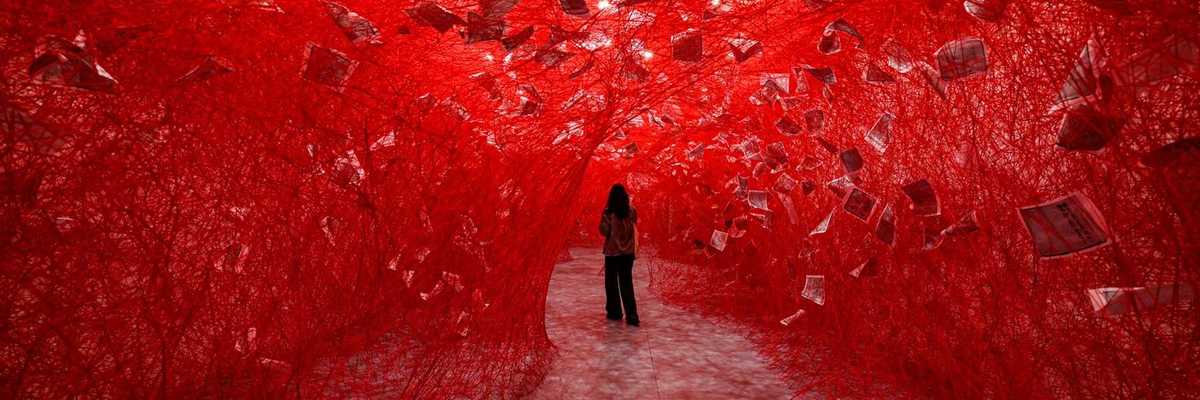 a figure standing in a tunnel of red woven yarn with pieces of paper suspended in it
