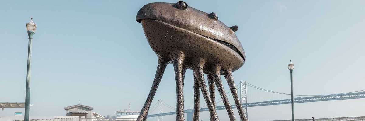 a large metal sculpture that looks like an alien with octopus legs in front of the Bay Bridge