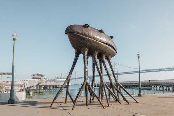 a large metal sculpture that looks like an alien with octopus legs in front of the Bay Bridge