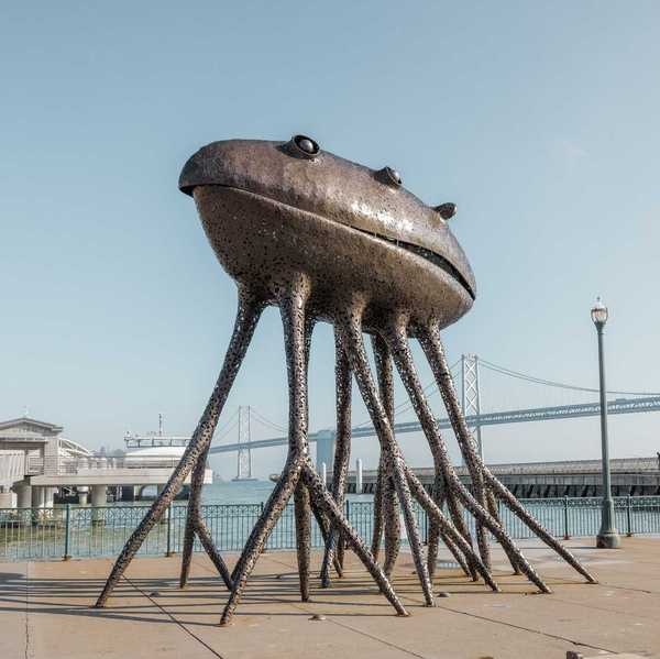 a large metal sculpture that looks like an alien with octopus legs in front of the Bay Bridge