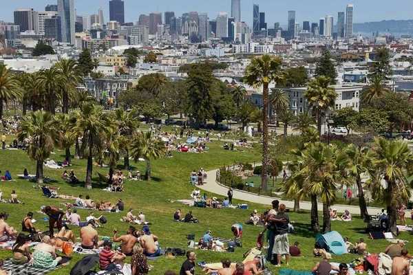 a park filled with people sitting on blankets with the SF city skyline behind