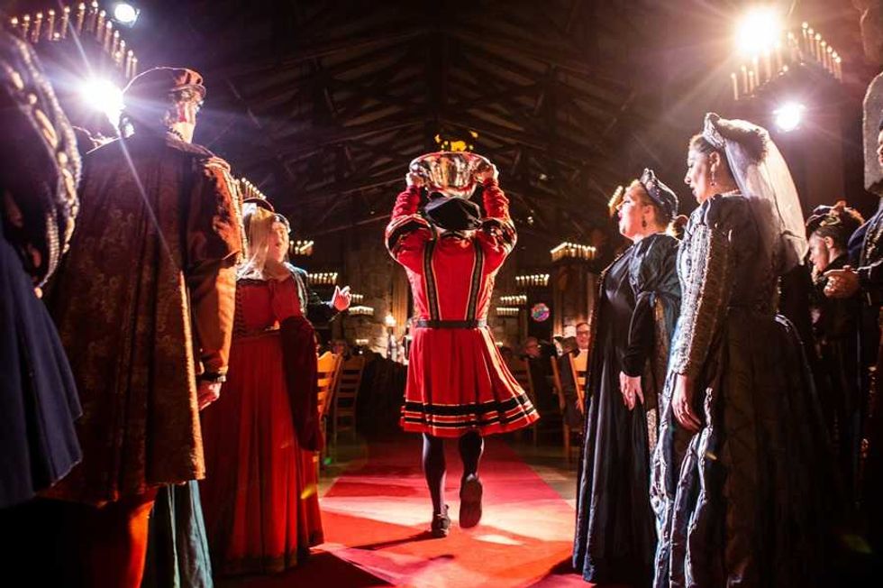 A person in medieval attire carries a large plate in a dimly lit banquet hall.
