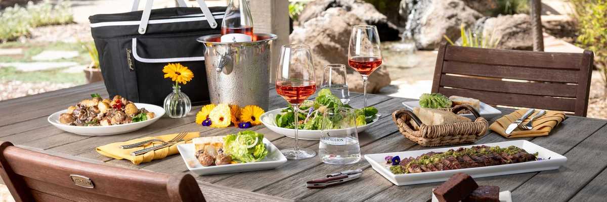 a picnic table with food and wine on top in a lush winery setting