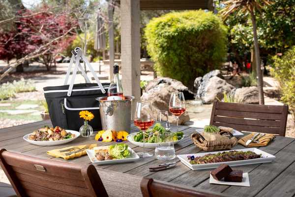a picnic table with food and wine on top in a lush winery setting