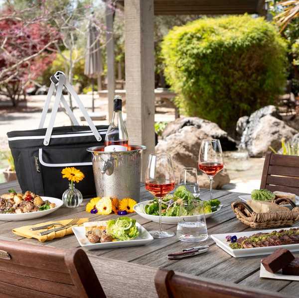 a picnic table with food and wine on top in a lush winery setting