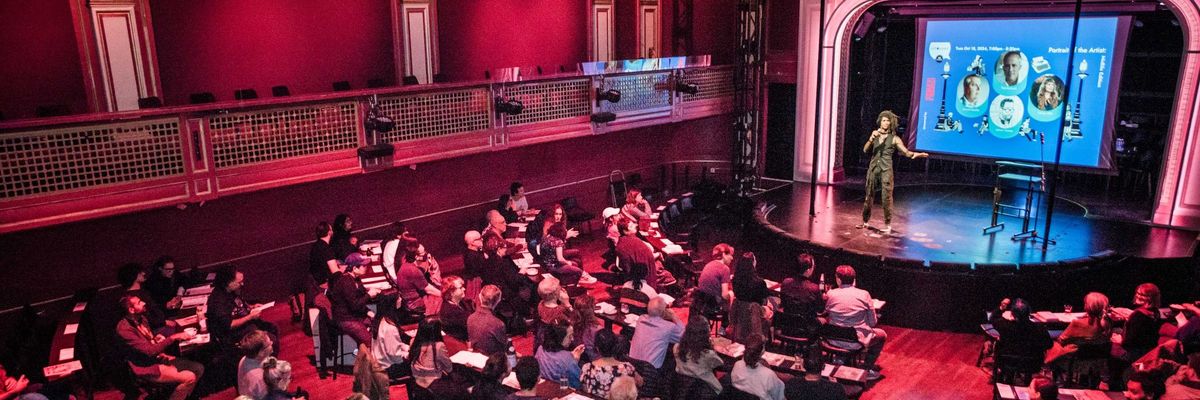 A speaker presents to a seated audience in a large, ornate theater.