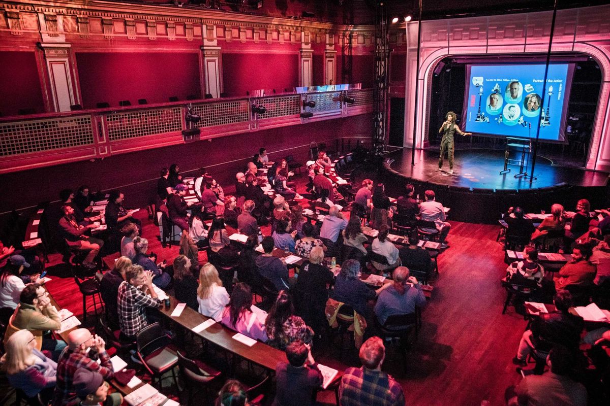 A speaker presents to a seated audience in a large, ornate theater.