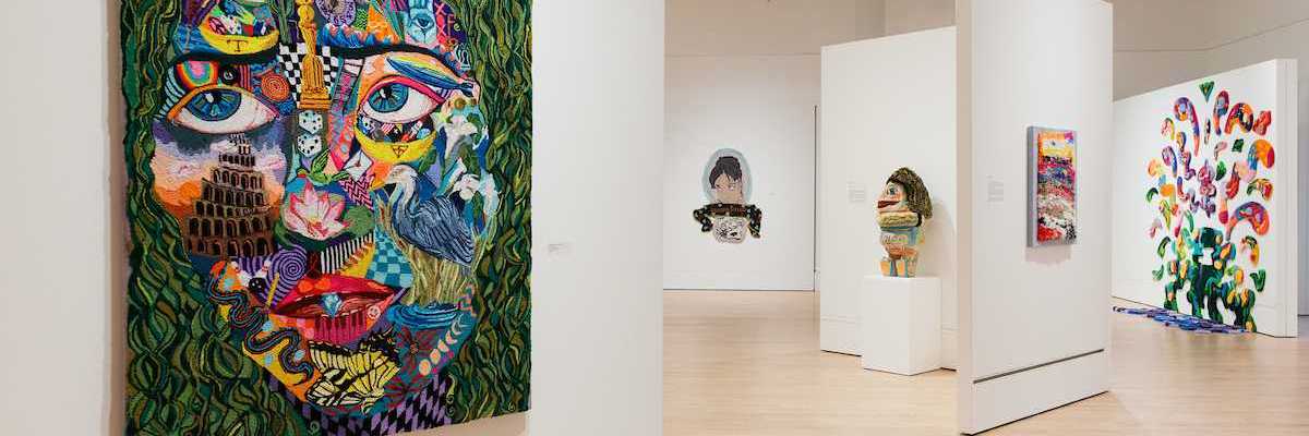 a tufted work of art that's a colorful face in a gallery with other works of art behind it