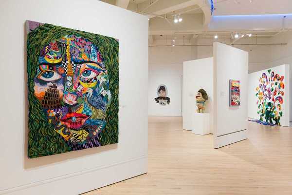 a tufted work of art that's a colorful face in a gallery with other works of art behind it