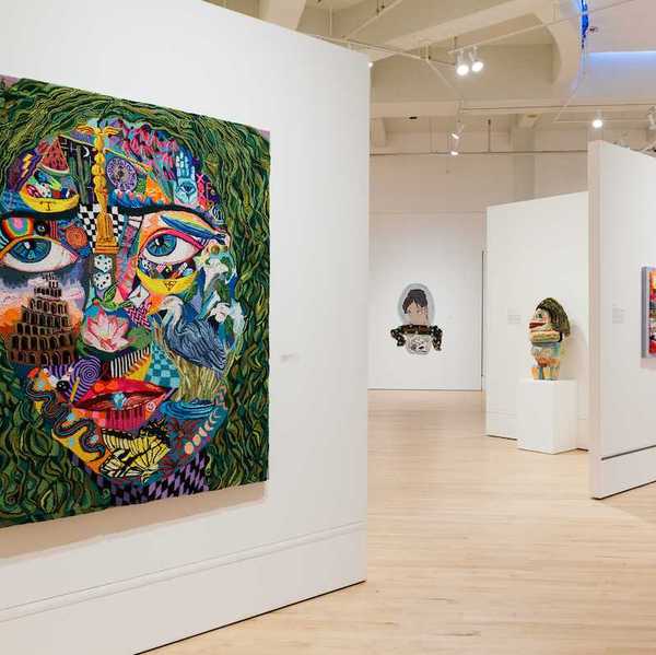 a tufted work of art that's a colorful face in a gallery with other works of art behind it