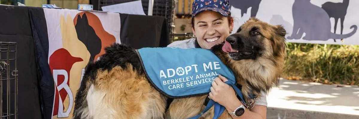 a woman in a hat and sneakers sitting with a German Shepherd mix wearing an "adopt me" vest