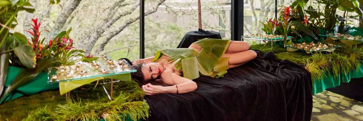 a woman lying naked on a banquet table covered in leaves with food around her and apples hanging from above