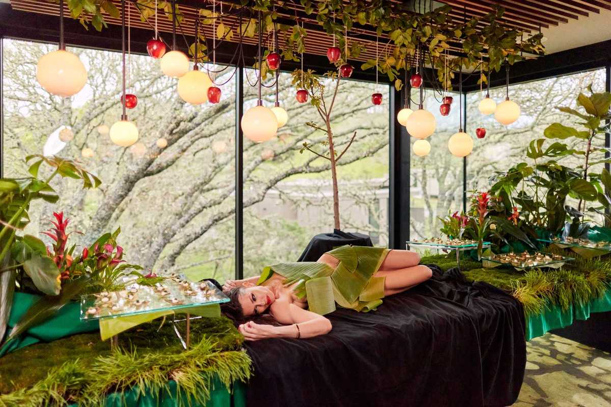 a woman lying naked on a banquet table covered in leaves with food around her and apples hanging from above