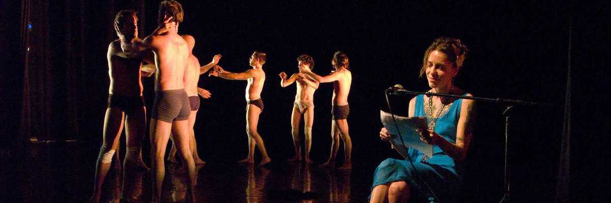 A woman sitting in a chair and reading on a dark stage with men dancing in black underwear behind her