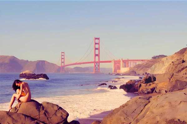 a woman sitting on a rock at the beach in front of the golden gate bridge