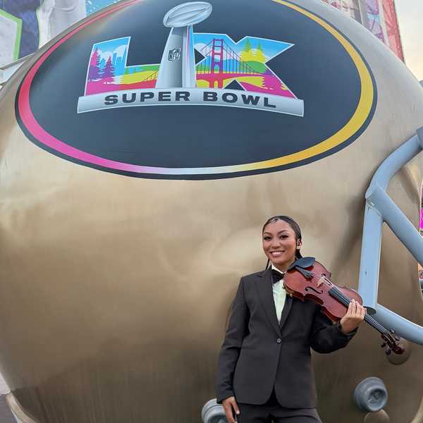 a woman with a violin standing in front of a giant football helmet that says LX Super Bowl