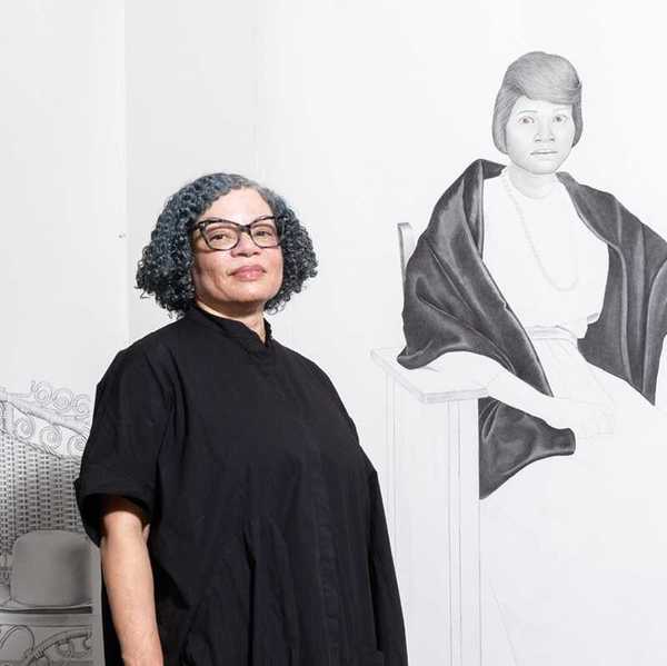 a woman with short curly hair and galsses in a black dress standing in front of two lareg pencil portraits