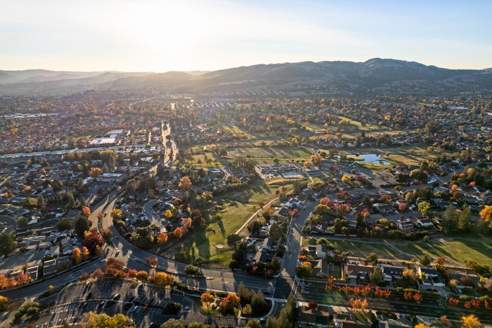 Aerial view of a suburban landscape at sunset with roads, houses, and autumn trees.