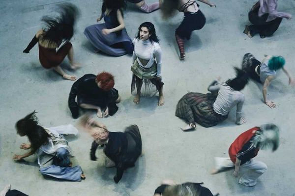 Aerial view of dancers in motion, surrounding one standing still, with blurred dynamic movements.