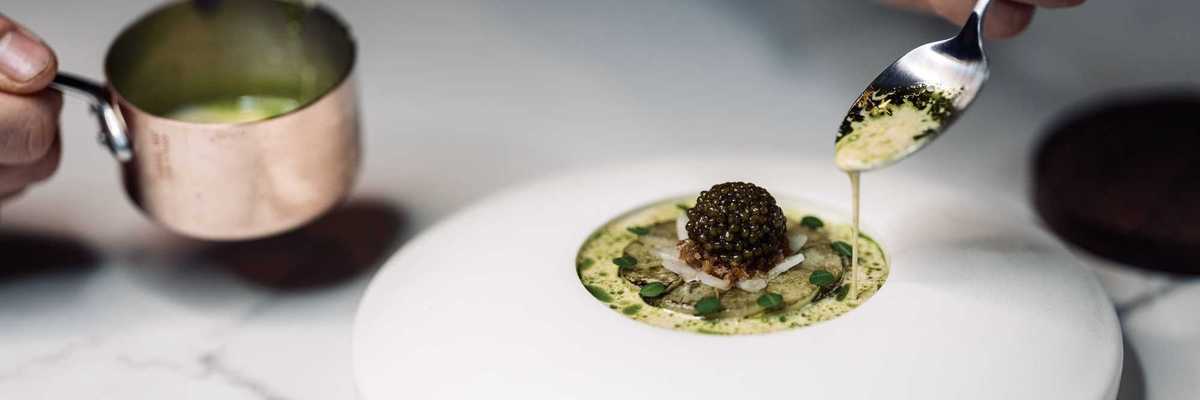 An elegant dish of food that includes a lump of caviar with a spoon drizzling sauce over it