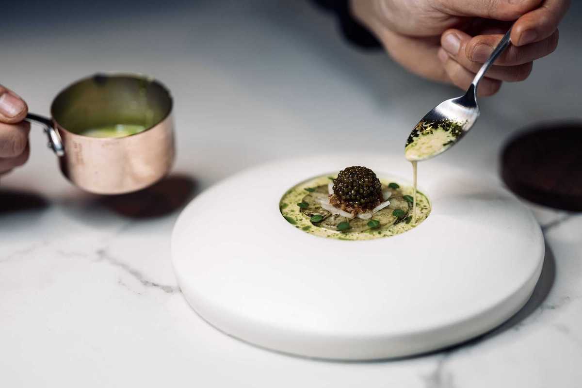 An elegant dish of food that includes a lump of caviar with a spoon drizzling sauce over it
