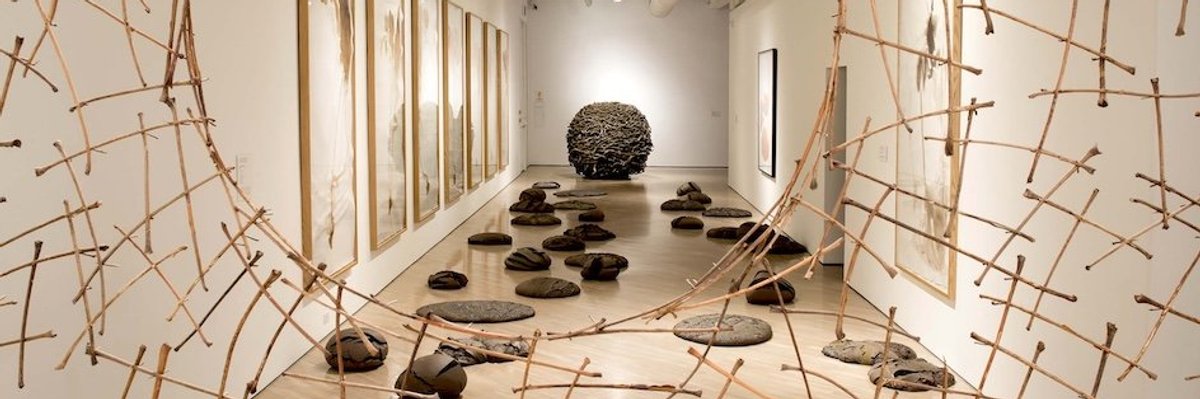 Art gallery with hanging wood sculpture, stones on floor, and framed artwork on walls.