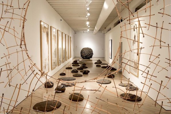 Art gallery with hanging wood sculpture, stones on floor, and framed artwork on walls.