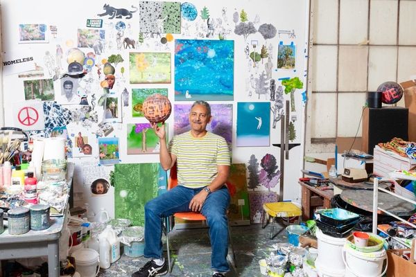 Artist in a colorful studio sits surrounded by artwork, holding a painted globe.
