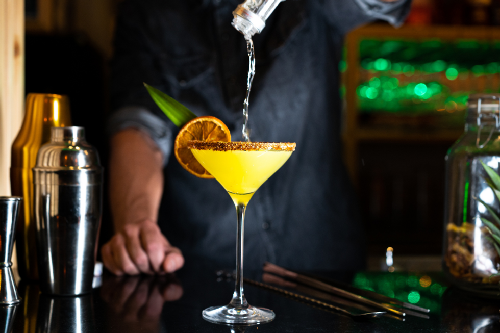 Bartender pouring a cocktail into a garnished glass, with shakers nearby.