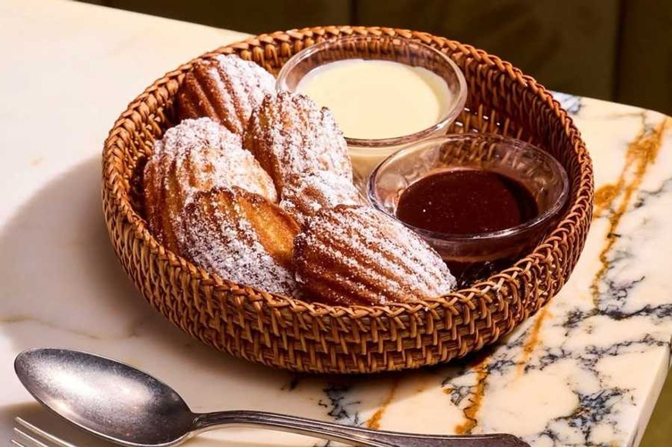 Basket of madeleines with powdered sugar, two dips, on a marble table with a spoon.
