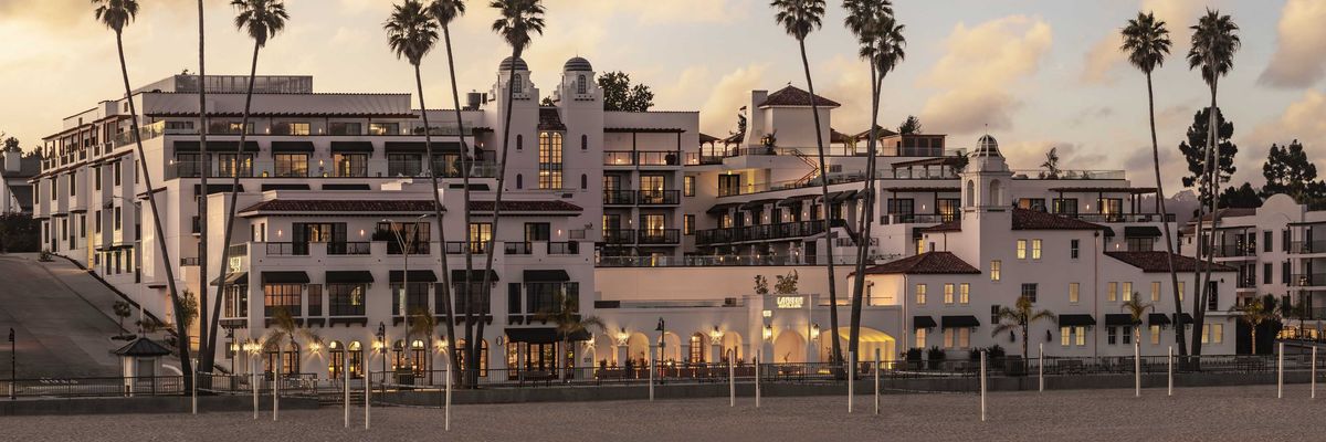 Beachfront hotel with palm trees at sunset, glowing warmly under a cloudy sky.