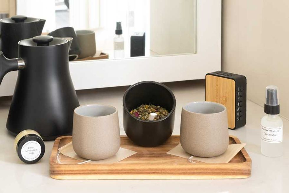 Black kettle, two mugs, herbs, and a spray bottle on a wooden tray by a mirror.