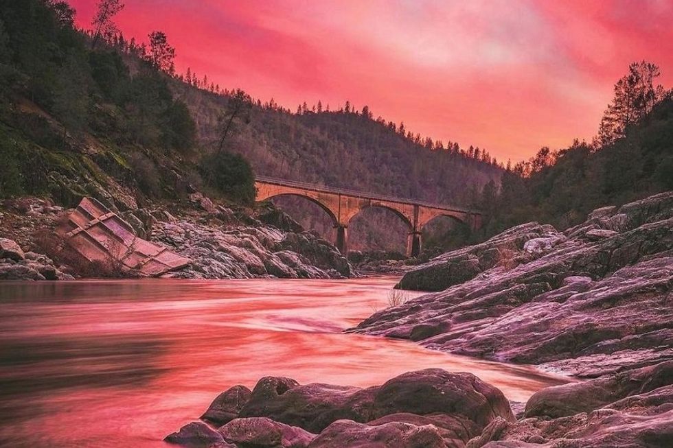 Bridge over a rocky river at sunset, pink sky and forested hills in the background.