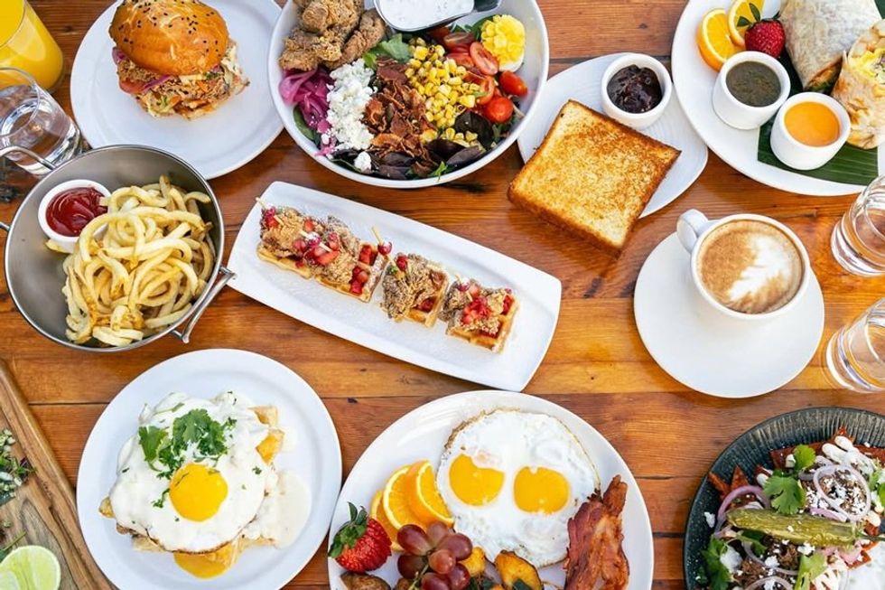 Brunch spread: burger, fries, salad, toast, coffee, eggs, fruit, and pastries on a wooden table.
