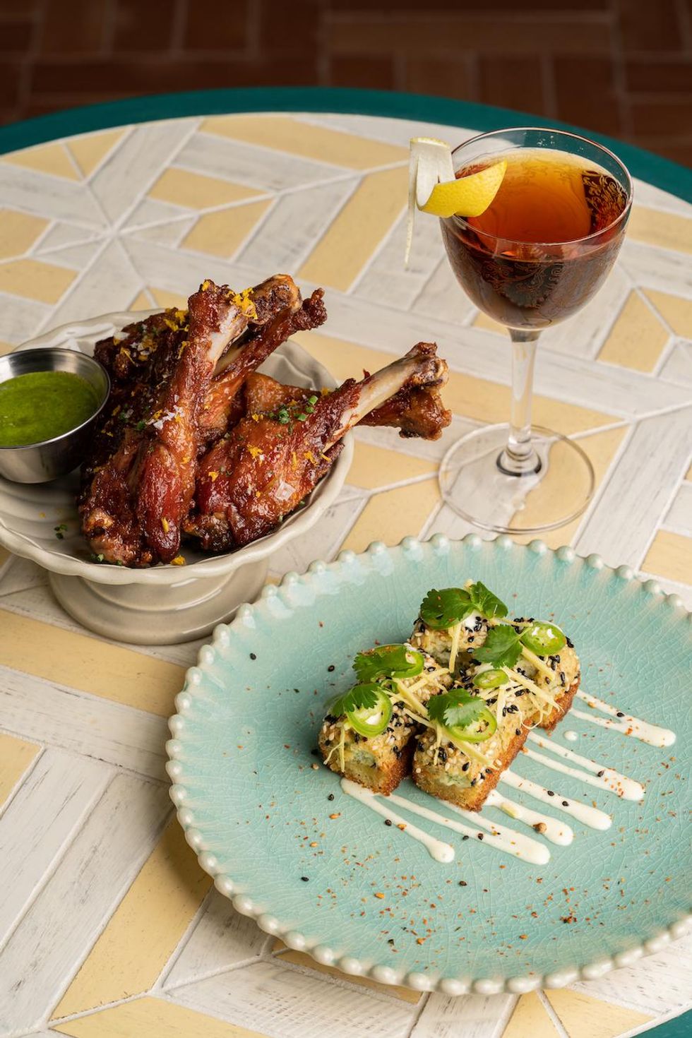 Chicken drumsticks, cocktail, and topped bread on a patterned table.