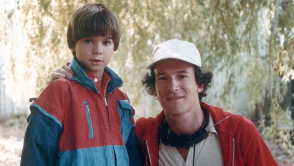 Child in a red and blue jacket with an adult in a cap outdoors.