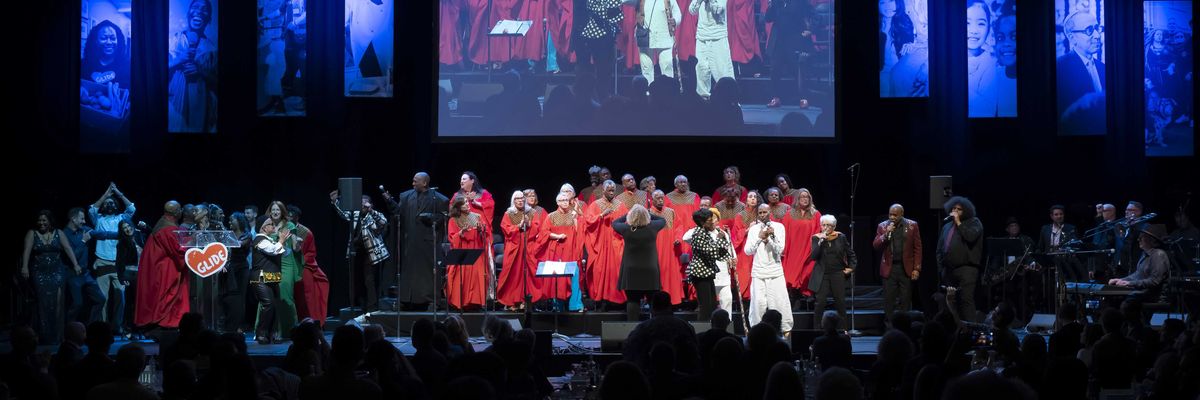 Choir performing on stage with singers in red robes and lit portraits in the background.