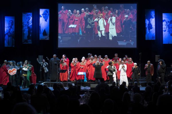 Choir performing on stage with singers in red robes and lit portraits in the background.