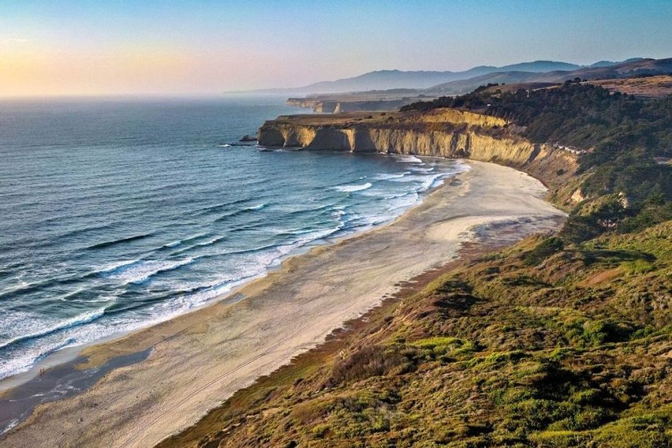 Coastal cliffs and sandy beach with ocean waves at sunset, surrounded by greenery.